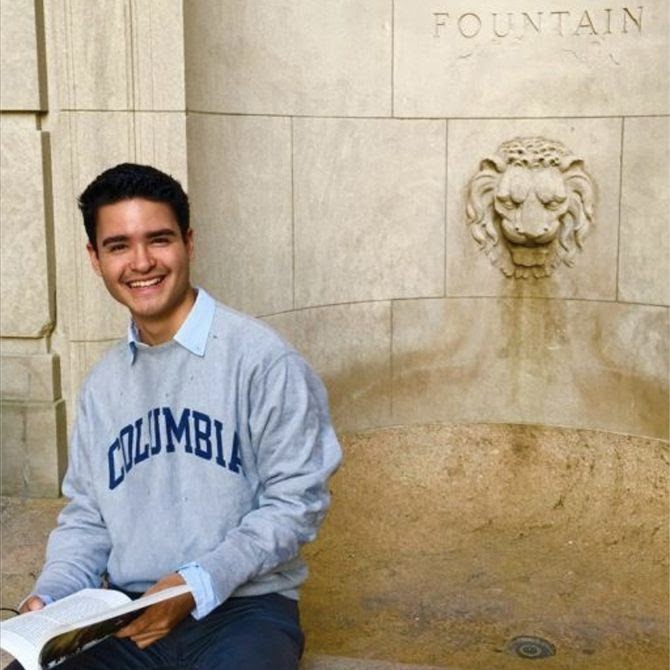 Santiago Tobar Potes, Columbia College Class of 2020, seated by Columbia Fountain in Columbia Sweater holding an open book on his lap